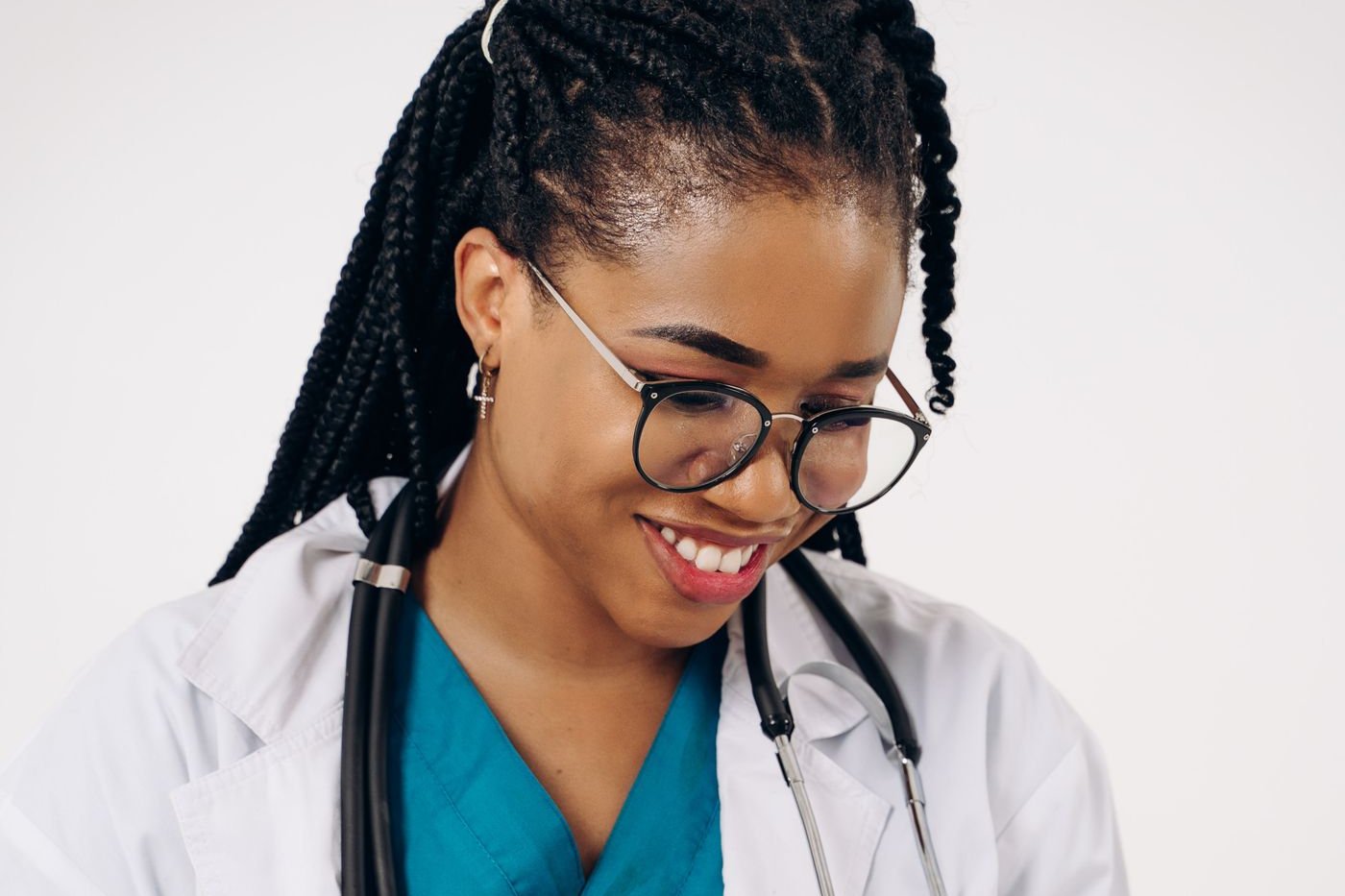 Smiling nurse in teal scrubs, a white coat and glasses with a stethoscope around her neck, looking down at her notes