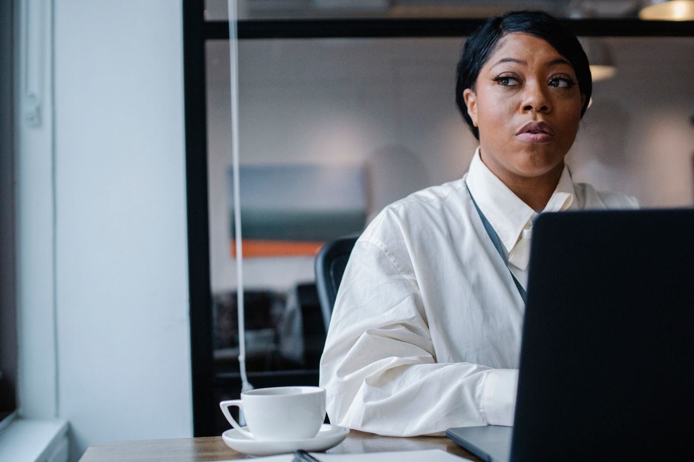 A professional woman in a white shirt sits at a laptop in a bright office, looking away in thought beside a cup of coffee