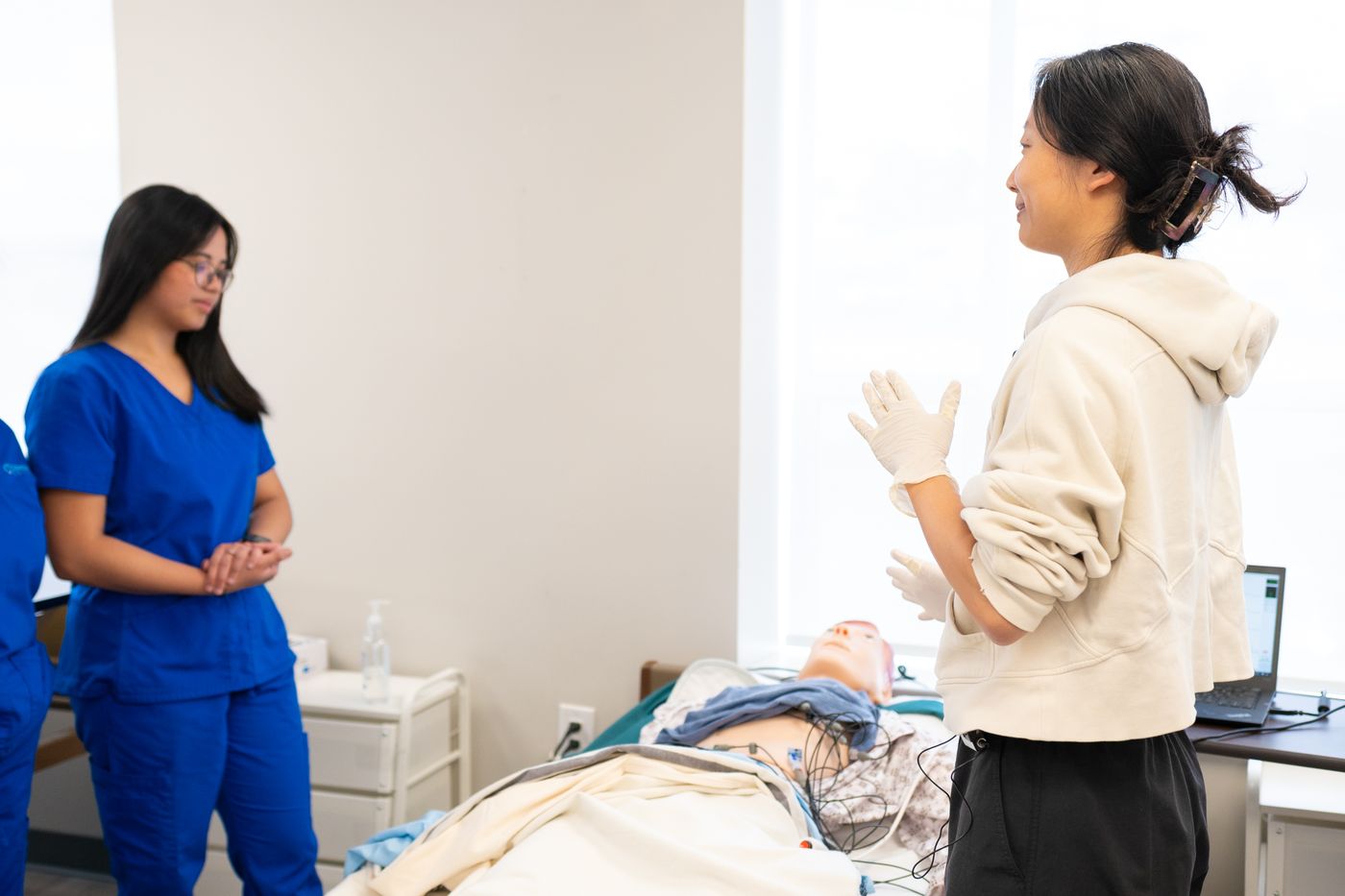 Nurse educator in a cream hoodie and gloves talking to a nursing student in blue scrubs beside a patient simulator manikin in a hospital bed in a bright skills lab