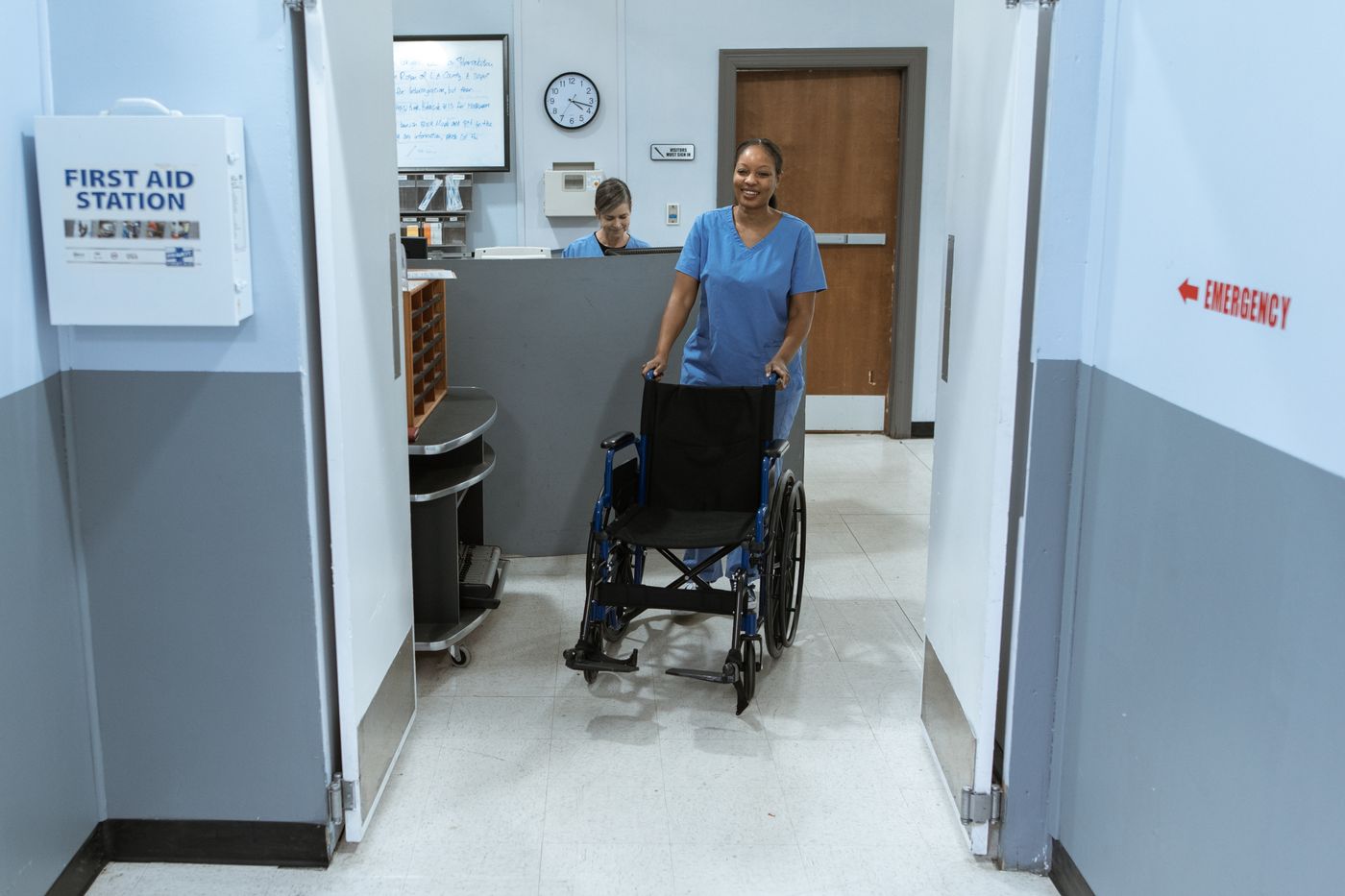 Smiling nurse in blue scrubs pushing an empty wheelchair through a hospital doorway past a First Aid Station sign, with a red Emergency arrow on the wall and a colleague at the desk behind her