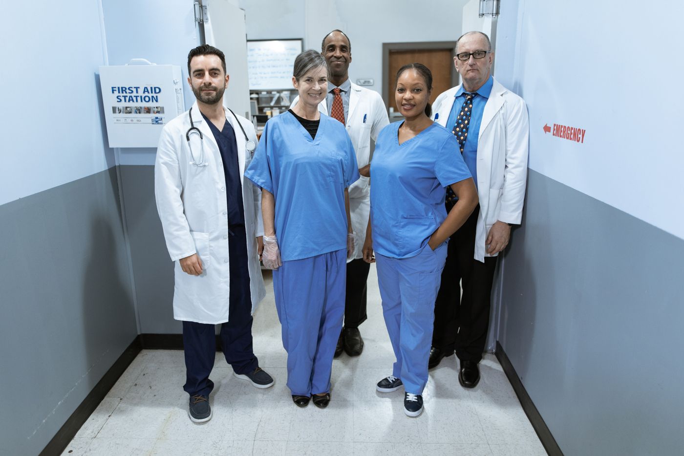 Five hospital staff standing together in a corridor beside a first aid station sign: two nurses in blue scrubs, one of them a Black woman, in front of three clinicians in white coats