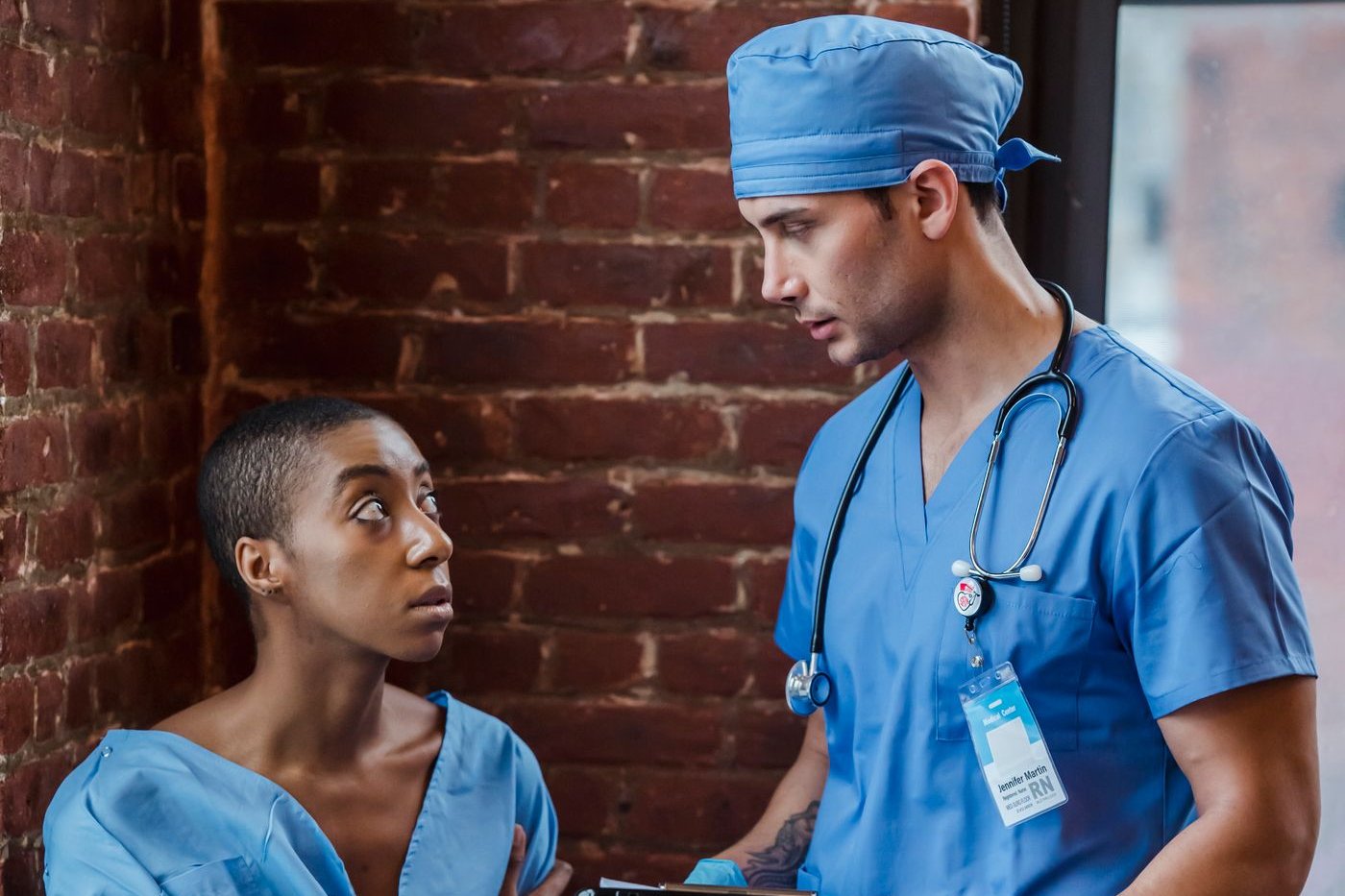 Woman in a patient gown looking up at a nurse in blue scrubs holding a clipboard during a simulated encounter