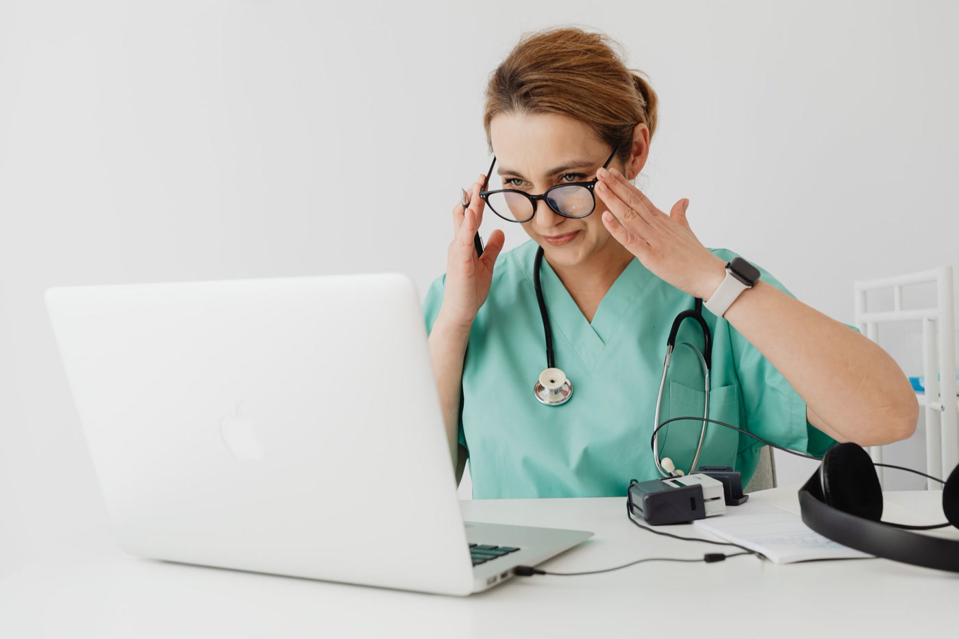 A nurse in teal scrubs and a stethoscope adjusting her glasses as she works through a course on her laptop