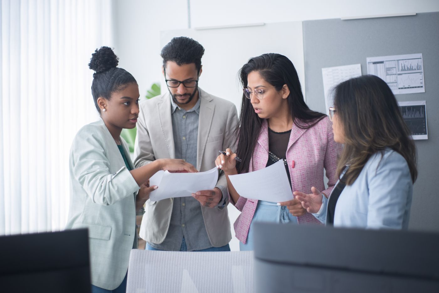 Four colleagues in business attire standing together in a bright office, reviewing printed documents, with charts pinned to a board behind them