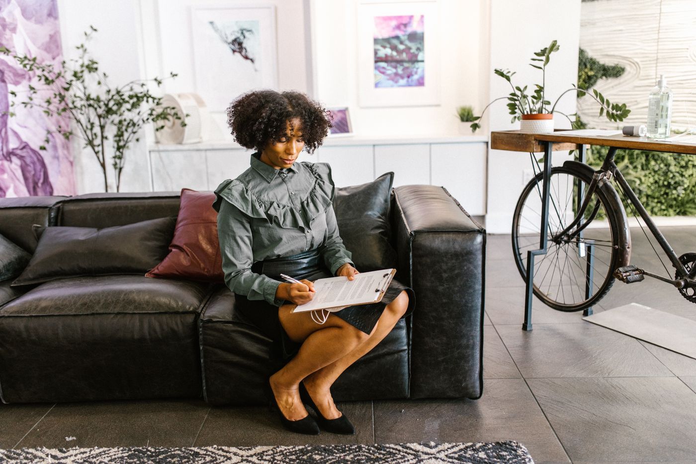 Woman with natural curly hair sitting on a black leather sofa in a bright living room, reading and marking a document on a clipboard