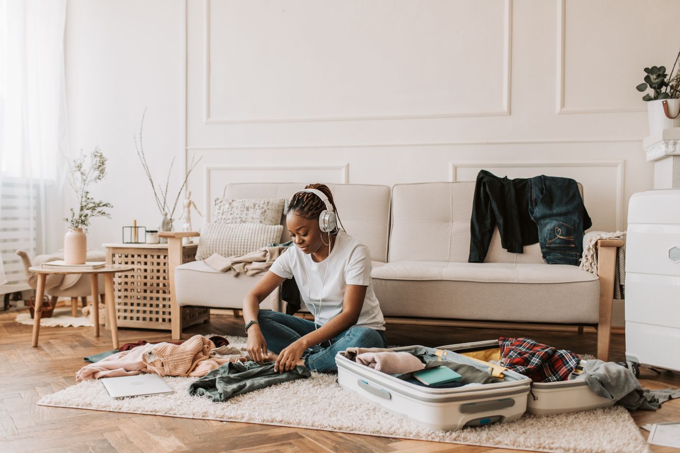 Young Black woman wearing headphones sits on a rug in a bright living room, folding clothes into an open white suitcase in front of a beige sofa
