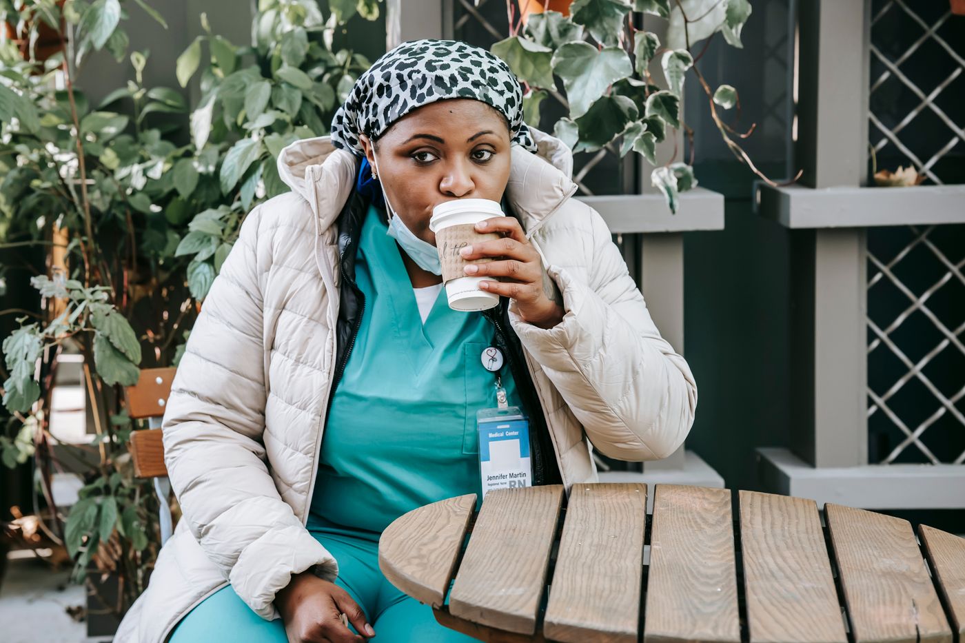Nurse in teal scrubs and a white puffer jacket sipping a takeaway coffee at an outdoor wooden table during a break, with leafy plants behind her