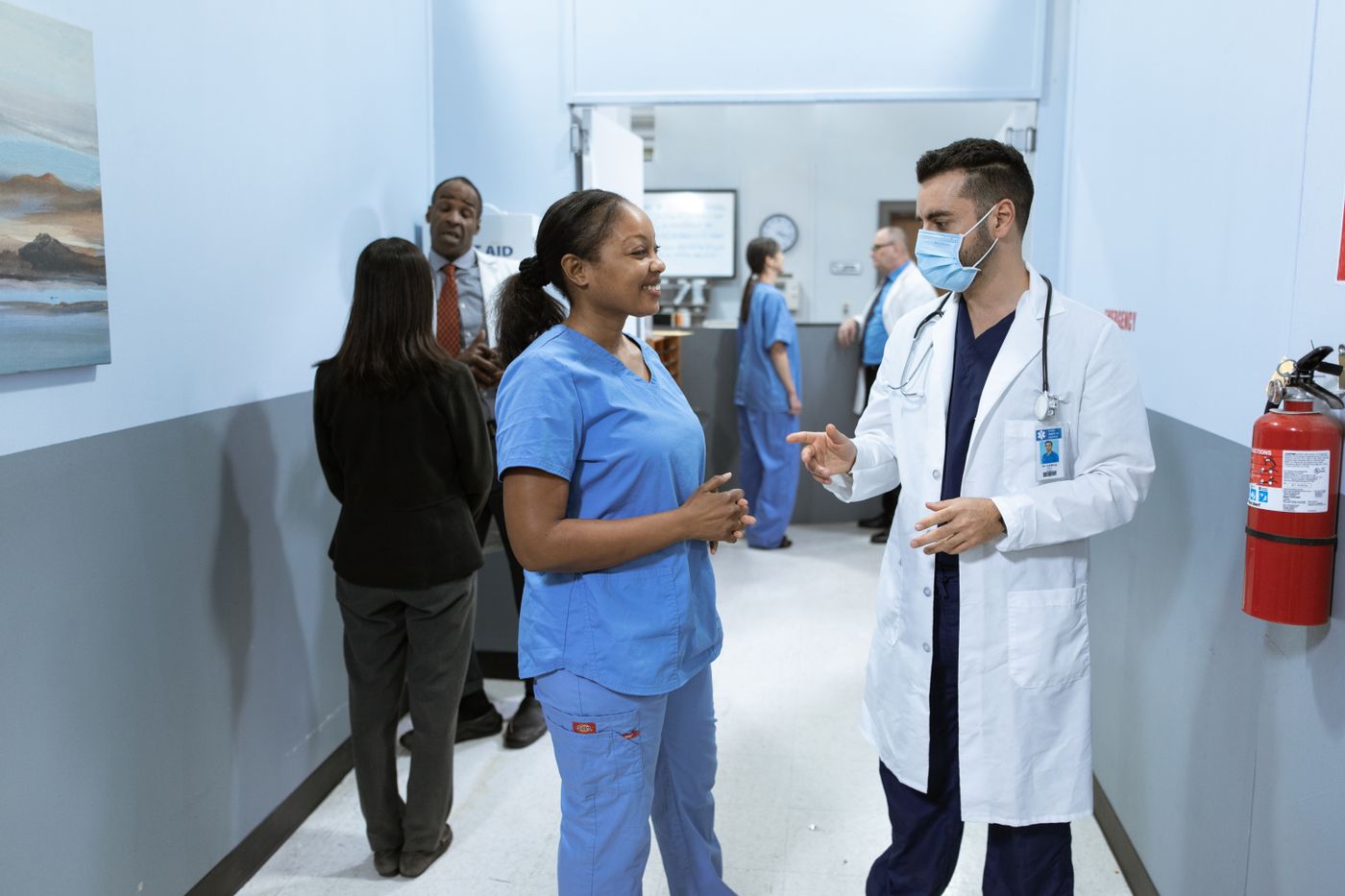 A smiling Black nurse in blue scrubs talks with a masked physician in a white coat in a hospital corridor, with colleagues talking near the nurses' station behind them